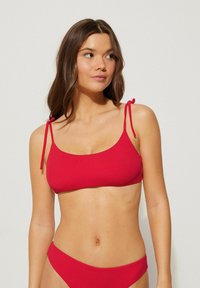 Červený bikiny top s tenkými nastavovacími ramínky a žebrovanou strukturou, spárovaný s odpovídajícími nízkými kalhotkami, které mají hladký design.