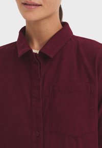 Camisa de pana burdeos con cuello puntiagudo, cierre frontal de botones y un único bolsillo en el pecho izquierdo. Detalle de tejido texturizado visible.