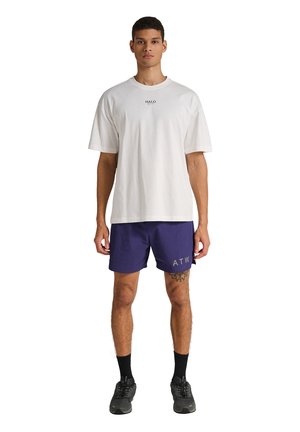 Mandlig model iført en hvid HALO T-shirt, mørkeblå ATW shorts, sorte sokker og sorte atletikskor, stående mod en hvid baggrund.