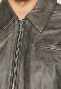 Närbild av en mörkgrå läderjacka med en silverfärgad dragkedja och en liten bröstficka, som bärs på en persons överkropp.