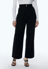 Pantalones de pierna ancha negros con una cintura alta, que presentan pliegues y dos botones metálicos. Hechos de una tela texturizada. Llevados con sandalias de tacón negras.