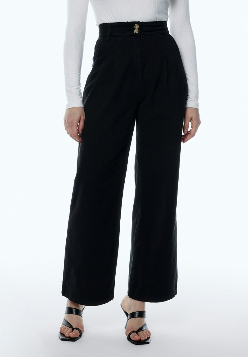 Pantalones de pierna ancha negros con una cintura alta, que presentan pliegues y dos botones metálicos. Hechos de una tela texturizada. Llevados con sandalias de tacón negras.