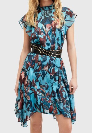 Kobieta ubrana w niebieską sukienkę w kwiatowy wzór z falbanami na rękawach i czarnym paskiem z ćwiekami w talii, stojąca na jednolitym tle.