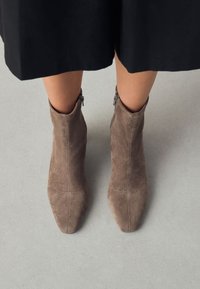 Stivaletti alla caviglia in camoscio marrone con punte appuntite, cerniere laterali e dettagli di cuciture sottili, su uno sfondo neutro.