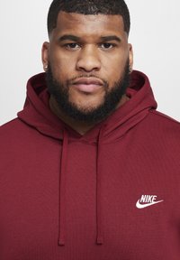 Bordeaux hoodie met een voortas, trekkoorden en een wit Nike-logo op de borst, gemaakt van een zachte katoenmix.