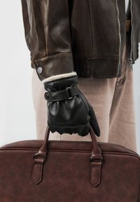 Sort læderhandske med manchet og spænde, båret på en hånd, der holder en brun lædertaske med dobbelte håndtag og tekstureret overflade. Brun læderjakke synlig.
