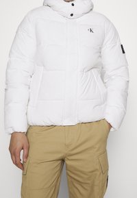 Giacca a vento bianca con collo alto, con un logo sul lato sinistro del petto e due tasche frontali. Indossata con pantaloni cargo color kaki.