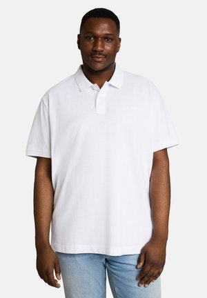 Mann steht und trägt ein schlichtes weißes kurzärmeliges Polohemd und hellblaue Jeans vor einem einfarbigen hellen Hintergrund.