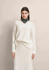 Rippstrick-Cremepullover mit langen Ärmeln und seitlichen Schlitzern, kombiniert mit einem schwarz-weißen geometrischen Musterschal um den Hals.