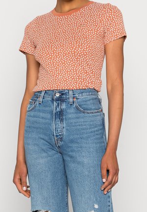 T-shirt à manches courtes de couleur rouille avec des motifs de fleurs blancs, associé à un short en denim bleu déchiré taille haute.