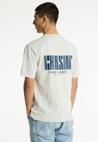 Camiseta de algodón color beige claro con un diseño gráfico azul en la parte posterior que presenta letras y texto en negrita, mangas cortas y un corte relajado.
