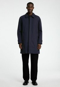 Temno moderna dolga bunda iz gladkega materiala, z ovratnikom, s centralnim gumbom za zapiranje in stranskimi žepi; v kombinaciji s črnimi hlačami.