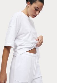 Weißes Baumwoll-T-Shirt mit kurzen Ärmeln, lockerer Passform und seitlichen Schlitzen, kombiniert mit weißen Jogginghosen mit elastischem Bund und frontalen Taschen.