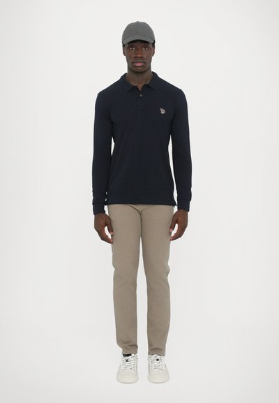 Pólo de manga longa azul marinho com colarinho, calças slim-fit bege, boné cinza e ténis brancos com atacadores padrões, em pé sobre um fundo simples.