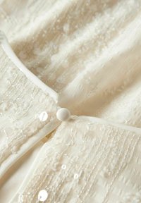 Gros plan d'un tissu de couleur crème avec des coutures texturées, des sequins et un bouton rond blanc fermant deux couches.