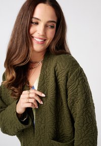 Olijfgroene gewatteerde jas met een tekstuurpatroon van bladeren, met een ontspannen pasvorm en een open voorkant. Laagjes van gouden kettingen en ringen zichtbaar.