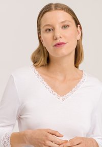 Giovane donna con capelli biondi lisci che indossa un top bianco con scollatura a V rifinito in pizzo, in posa contro uno sfondo chiaro e neutro.