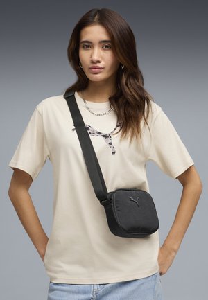 Jonge vrouw met lang haar, gekleed in een beige Puma t-shirt en een zwarte crossbody tas met een Puma-logo tegen een grijze achtergrond.