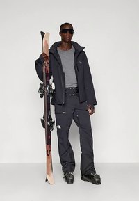 Skiausrüstung mit schwarzer Jacke und Hose, grauem Oberteil und schwarzen Skischuhen. Hält ein Paar Skier mit Bindungen und heller Holzsohle.