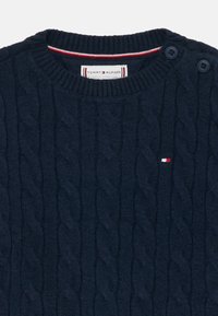 Mornarsko plavi pulover s pletenicom, s rebrastim ovratnikom, dva gumba na ramenu i malim logotipom Tommy Hilfigera na prsima.