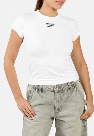 Vrouw draagt een aansluitend wit Reebok T-shirt en grijze hooggetailleerde jeans met handen in de zakken.