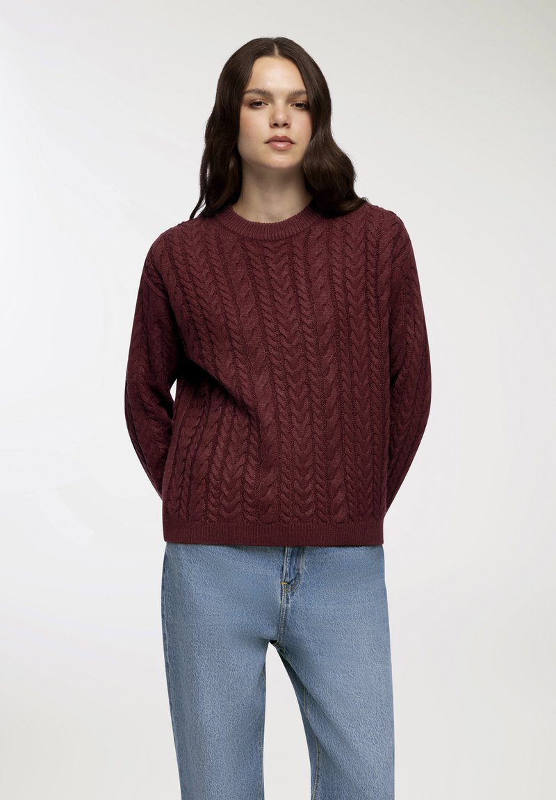 Maglione in burgundy lavorato a maglia con motivo a treccia, colletto rotondo e polsini a coste, abbinato a jeans blu chiaro.