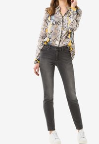 Camisa de estampado de serpiente parcialmente desabrochada en tonos marrón, gris, azul y amarillo; combinada con jeans skinny grises y zapatillas blancas.