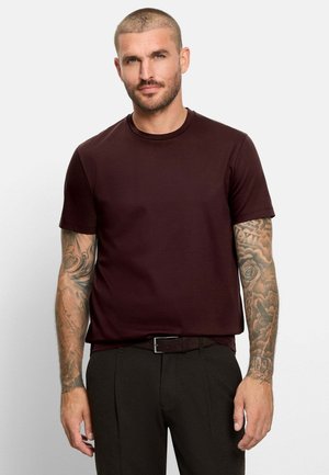 Uomo con capelli corti e tatuaggi sulle maniche, che indossa una maglietta marrone scuro e pantaloni neri, in piedi davanti a uno sfondo chiaro e uniforme.