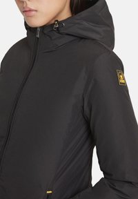 Giacca impermeabile nera con cappuccio, dotata di una zip frontale e di una toppa logo gialla sul braccio sinistro. Tessuto liscio e opaco.