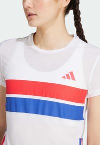 Camiseta deportiva blanca con franjas horizontales rojas y azules, fabricada con tejido transpirable. Presenta un cuello redondo y el logo de Adidas en el pecho.