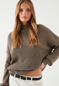 Brązowy sweter z prążkowanej dzianiny z wysokim kołnierzem i opadającymi ramionami, zestawiony z czarnym paskiem i białymi spodniami. Teksturowana tkanina, luźny krój.