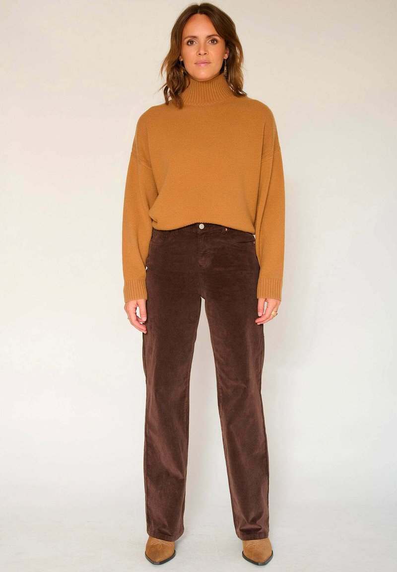 Pantalon en velours côtelé marron et pull en tricot beige à col roulé, avec une coupe décontractée. Les chaussures sont de couleur brun clair avec un bout pointu.