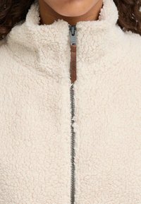 Giacca in pile con zip color crema e colletto alto, dotata di una zip scura e un pull tab in pelle per la chiusura. Tessuto morbido e testurizzato.