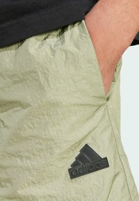 Shorts in tessuto crinkle di un verde oliva chiaro, con un logo Adidas ricamato in nero sul lato sinistro e apertura delle tasche inclinata.