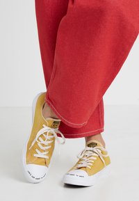 Baskets en toile jaune avec semelles et lacets en caoutchouc blancs, arborant l'inscription « LIFE'S TOO SHORT TO WASTE » sur l'avant. Portées avec un pantalon rouge.