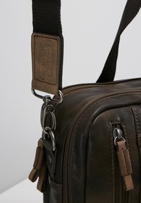 Braune Lederhandtasche mit strukturierten Oberfläche, ausgestattet mit einem schwarzen Tragegurt, silbernen Hardware und kleinen Reißverschlussfächern mit Lederzugbändern.
