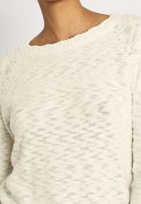 Ljusbeige stickad tröja med vid halsringning, strukturerat mönster och ribbade detaljer längs halsringningen och ärmarnas avslut.