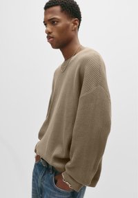 Jongeman in een beige wafelgebreide trui, zilveren ketting, en blauwe spijkerbroek, staand met zijn handen in de zakken tegen een witte achtergrond.