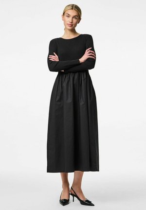 Black long-sleeve dress with a fitted top and a flared, gathered skirt. Textured fabric, mid-calf length, paired with black heeled shoes.
