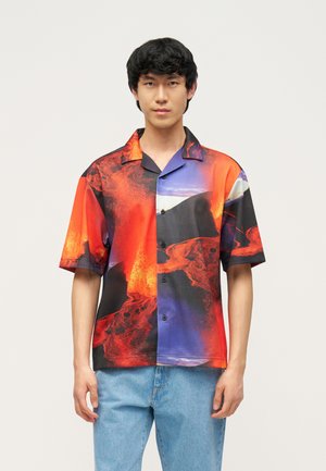 Jeune homme portant une chemise à manches courtes vibrante avec un motif abstrait de lave rouge, noire et bleue, associée à un jean bleu clair.