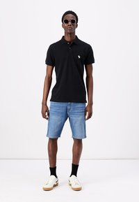 Črna polo majica s ovratnikom, oznaka logotipa, v kombinaciji z svetlo modrimi denim kratkimi hlačami. Obuta v bele superge in črne nogavice, dodatki vključujejo sončna očala.