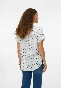 Chemise blanche à manches courtes avec des pois navy, à la coupe décontractée, ourlet arrondi et détail de poche à l'arrière. Associée à un pantalon en denim à jambes larges.