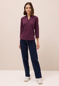 Bordeauxrotes Langarm-Polohemd mit Kragen und Knopfverschluss, kombiniert mit dunkelblauen geraden Jeans und weißen Sneakers.