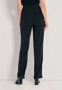 Persoon in hoge taille zwarte gestructureerde wijde pijpenbroek en zwarte hakken, staande tegen een effen lichte achtergrond.