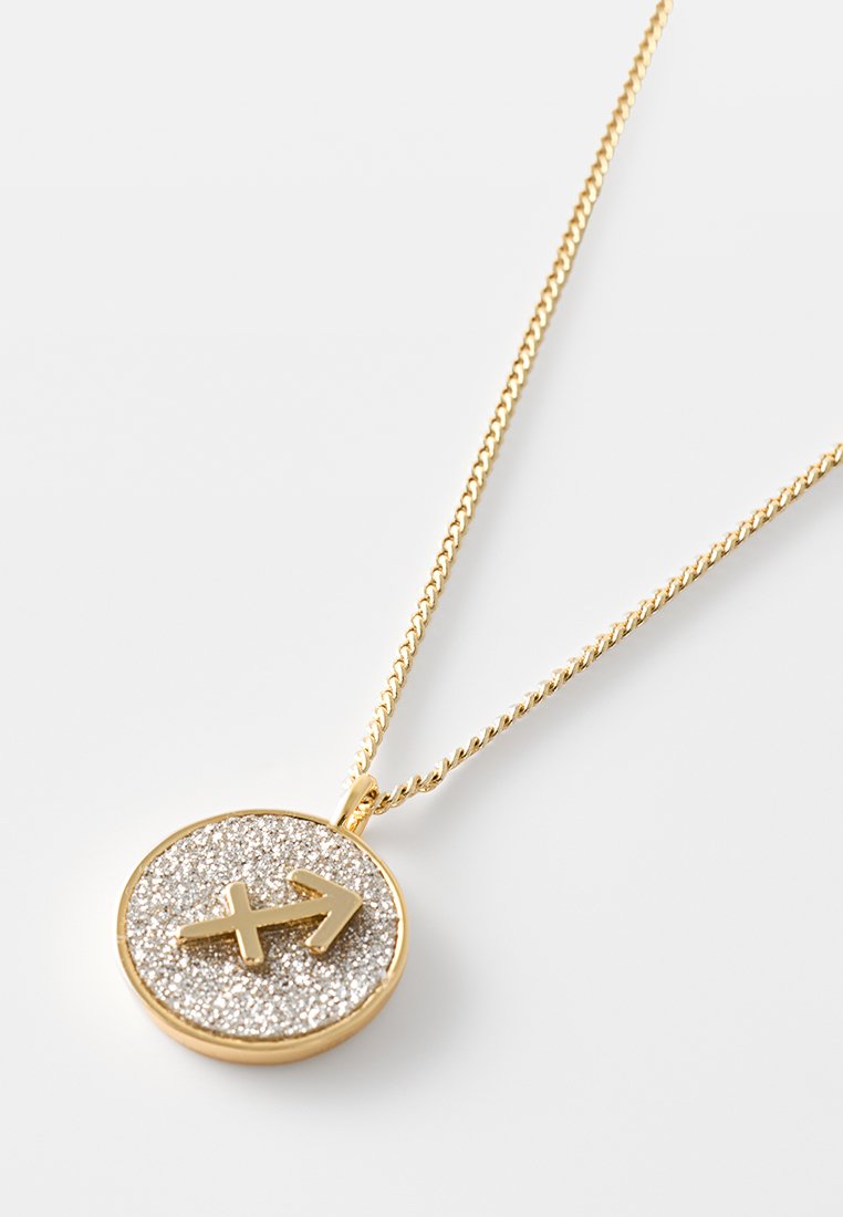 Złoty naszyjnik z okrągłym wisiorkiem; wisiorek ma błyszczącą srebrną powierzchnię z wytłoczonym wzorem strzały. Łańcuszek jest skręcony i delikatny.