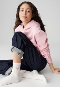 Roze sweatshirt, donkere spijkerbroek met opgerolde pijpen en witte sokken met roze hartjespatroon. Model zit op een neutrale achtergrond.