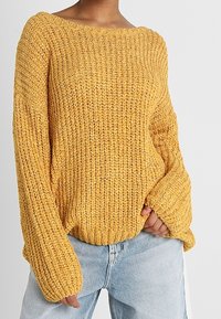 Mustard yellow knitted sweater with a wide neckline, loose long sleeves, and ribbed cuffs, paired with light blue high-waisted jeans.