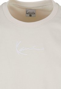Kremfarget bomulls t-skjorte med ribbet rund hals og et brodert logo i hvitt. Merket har en kursivert "Kane" og størrelse "M."