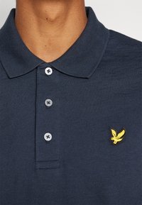 Mornarska polo majica z žametnim ovratnikom in tremi gumbi, ki ima na prsih izvezen majhen rumen orel. Mehka tekstura.