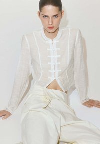 Jeune femme aux cheveux coiffés en arrière porte un haut blanc texturé avec des fermetures grenouille et un pantalon ivoire taille haute, assise devant un fond blanc.